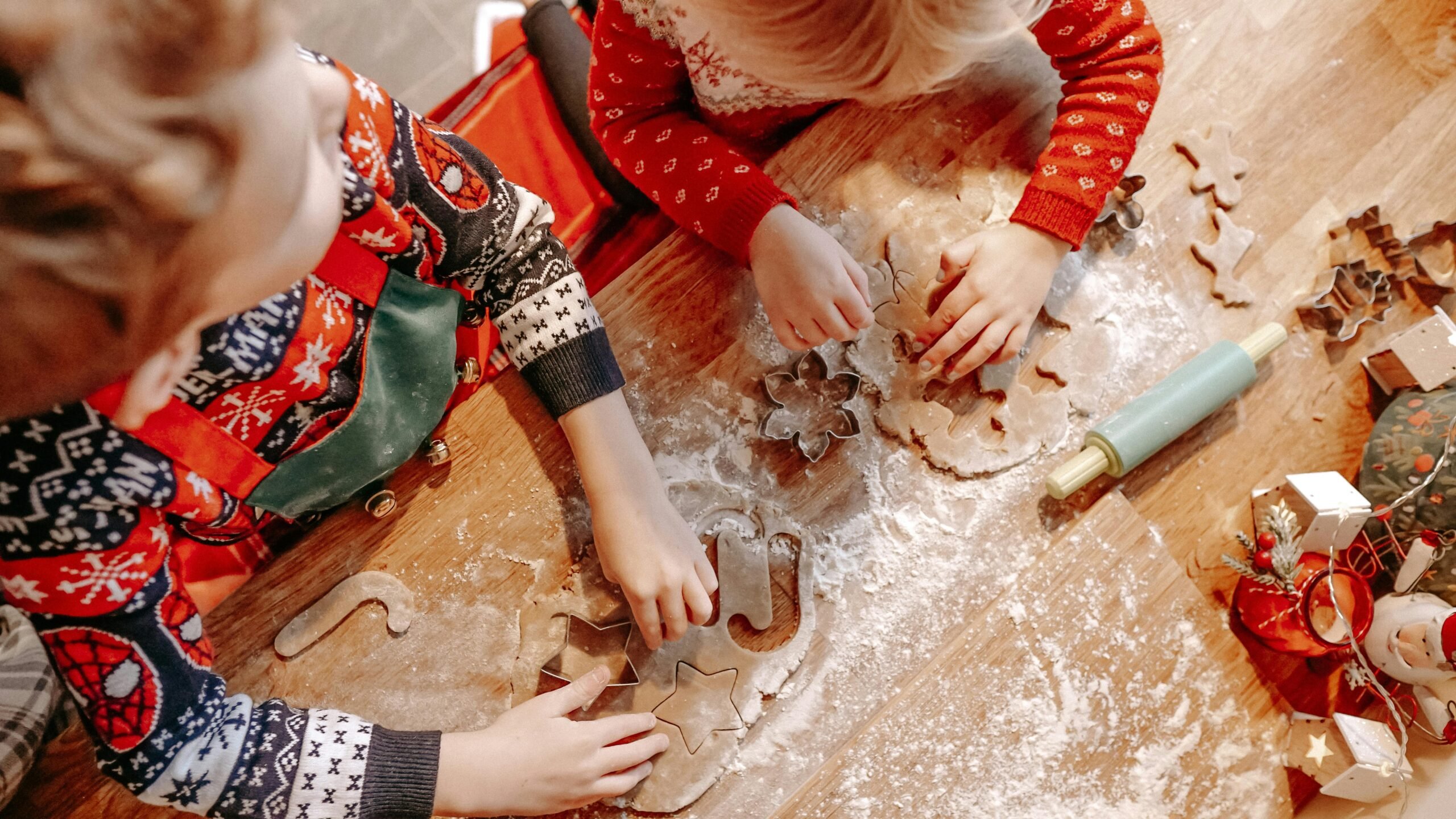Kids in festive sweaters make holiday cookies with cutter shapes on a wooden table.